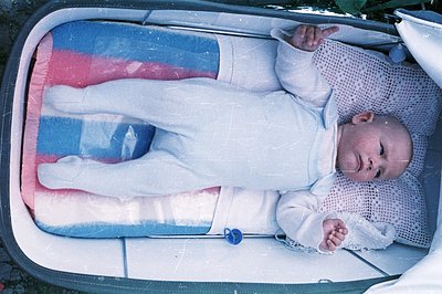 Infant rests within a vintage, striped stroller. Blue and pink horizontal lines adorn a soft blanket. The baby wears a white ...