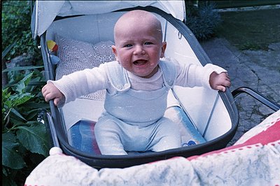 Infant in a vintage, metal-framed stroller, wearing light blue overalls and a long-sleeved top. Close-up view highlights a ha...