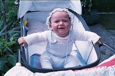 Infant in a stroller, wearing a bonnet & light blue overalls. The vintage stroller features a woven seat and metal frame. Out...