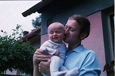 A man holds a baby close, smiling. The scene captures a tender moment, likely a family portrait. Architectural details sugges...