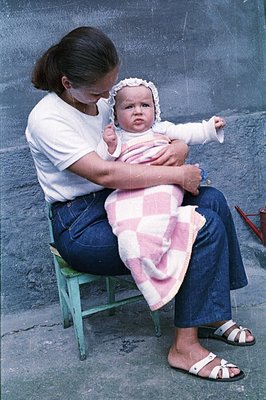 A young woman in a white tee and dark jeans sits on a green stool, cradling a baby bundled in white and pink. The baby wears ...