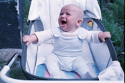 Infant in a stroller, expressing distress or excitement with an open mouth and raised arms. Wearing a white romper with butto...