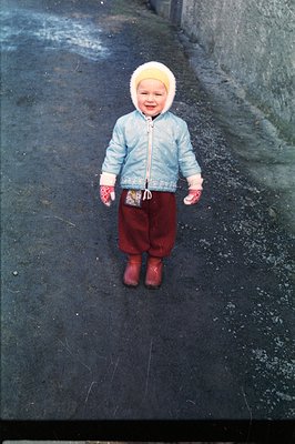 A young child stands on a paved road, bundled in a light blue parka, white fur hat, red snow pants, and red boots. The backgr...