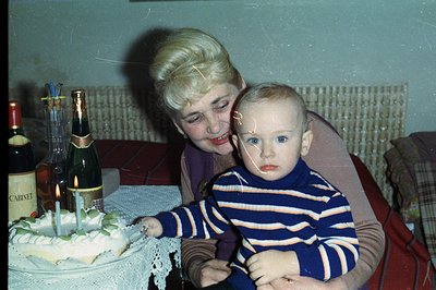 A mother and young child celebrate a birthday, evidenced by a layered cake with candles and champagne. The mother has a disti...