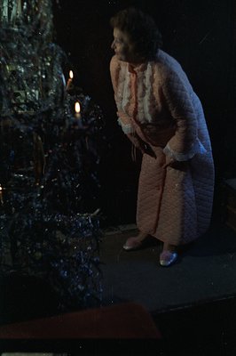 A woman in a long, quilted, peach-colored robe stands near a Christmas tree with lit candles. The warm glow illuminates her f...