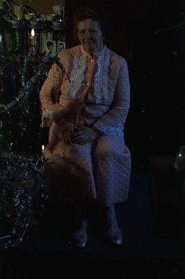 A woman sits posed in a pink quilted dress suit with ruffled collar, holding a decorative fan. A Christmas tree with vintage ...