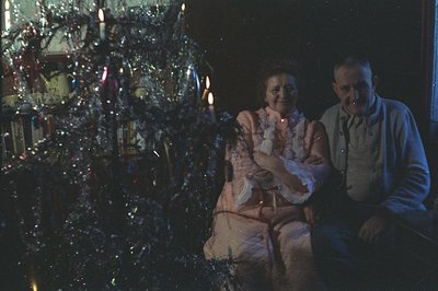 A couple sits near a brightly decorated Christmas tree with candles and string lights, photographed in color. The woman wears...