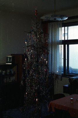Interior view of a warmly lit room featuring a tinsel-covered Christmas tree adorned with vintage ornaments. A red tablecloth...
