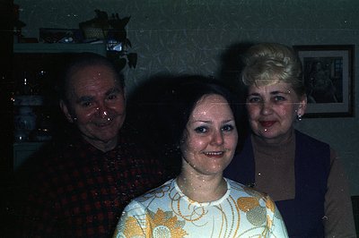 Three people pose indoors, likely a family portrait. The woman in the center wears a patterned yellow top, flanked by a man i...