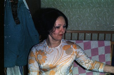 A woman with dark hair and prominent eye makeup sits on a bed with a pink and white quilted coverlet. She wears a cream-color...