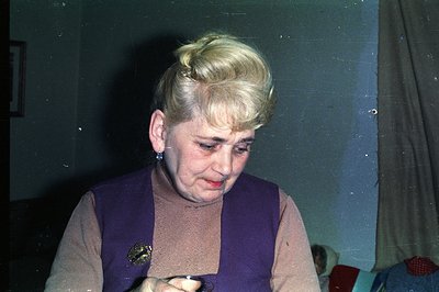 Close-up of a woman with blonde, stylized hair, wearing a purple vest over a beige sweater. She appears to be concentrating o...