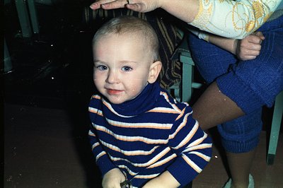 A toddler with light hair and blue eyes smiles directly at the camera, wearing a striped blue and orange turtleneck sweater. ...