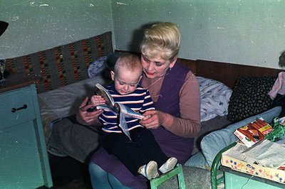 A young boy sits on a woman’s lap, both intently observing a magazine. The woman, with a distinctive 1960s hairstyle, wears a...