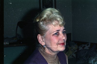 Close-up portrait of a woman with styled blonde hair, red lipstick, and pearl earrings. She wears a purple turtleneck sweater...