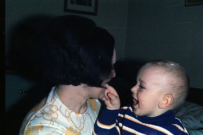 A young boy, likely a toddler, stands facing an adult woman, appearing to playfully cover her mouth with his hands. The woman...
