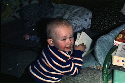 A young, bald boy sits on a bed, clutching a book, displaying a wide smile. He wears a dark blue and orange striped sweater. ...