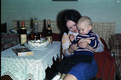 A woman with dark hair sits on a bench, cradling a baby, beside a table set with refreshments. A cake with lit candles, bottl...