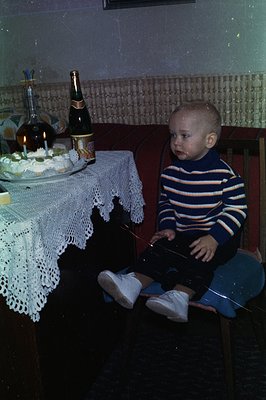 A young child sits attentively near a celebratory table laden with a cake, champagne, and a decanter. The child wears a strip...
