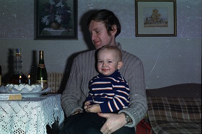 A man holds a baby in his lap, both smiling, within a warmly lit, likely domestic setting. The table displays a bottle of cha...