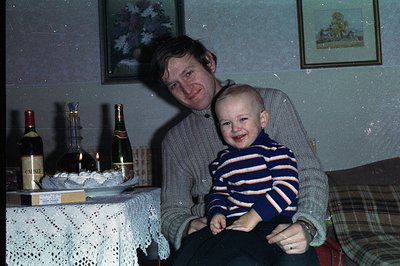 A smiling man in a grey sweater holds a young child wearing a striped sweater, posed indoors. A table with wine bottles, cand...