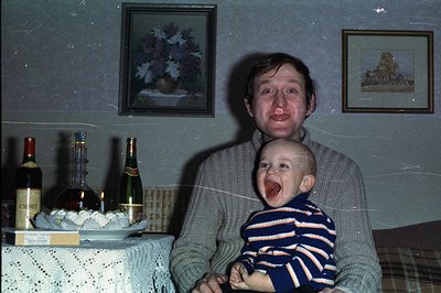 Man in a turtleneck sweater holds a baby who is mid-cry. Festive table with wine, champagne, and sweets is visible. Home inte...