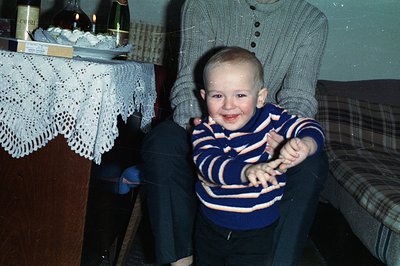 A smiling toddler, likely in the 1970s, wears a patterned sweater and dark trousers, playfully posed on the lap of an adult, ...