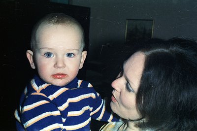 A young boy with striking blue eyes gazes directly at the camera, wearing a blue and orange striped sweater. A woman, partial...
