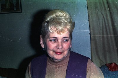 Close-up portrait of a mature woman with a distinctive, voluminous blonde hairstyle. She wears a beige turtleneck sweater and...