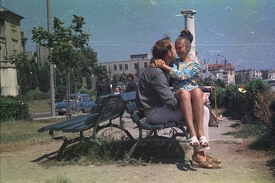 A young couple shares a moment on a park bench. The woman, in a floral print dress, sits facing the camera, while the man emb...