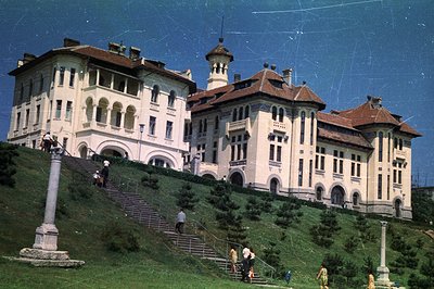 A grand, multi-story building with a red-tiled roof and ornate facade rises up a steep, verdant hillside. Several people asce...