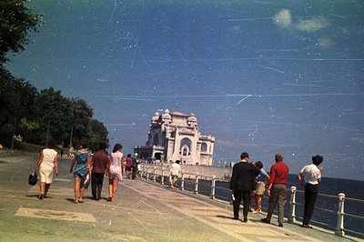 Seaside promenade with ornate architecture, likely a casino or pavilion. People stroll along the walkway, suggesting a leisur...