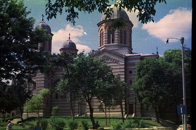 A grand, Neo-Byzantine cathedral dominates the view, featuring three prominent domes and intricate stonework detailing. Lush ...