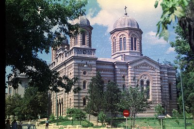 Exterior view of a grand, ornate stone cathedral with visible domes and a cross atop. Likely Eastern Orthodox, featuring elab...