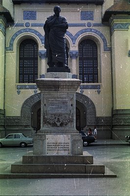 A bronze statue of a man in classical robes stands atop a large stone pedestal inscribed with text in Romanian. The backgroun...