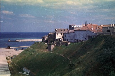 Coastal townscape showing hillside homes with terracotta roofs overlooking a sandy beach and the Black Sea. Appears to be a m...