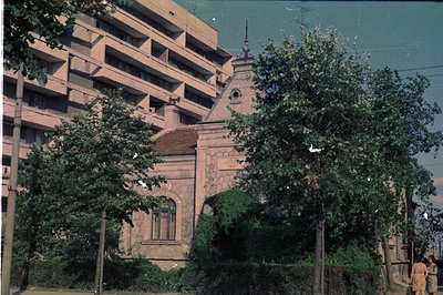 Exterior view of a detailed stone building, possibly a gatehouse or small public building, in front of a block of Brutalist a...