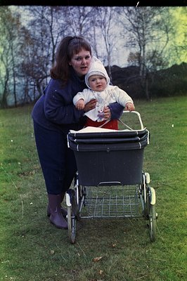 A young woman in a dark turtleneck and slacks stands next to a vintage pram, holding a baby bundled in a white snowsuit and b...