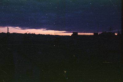 Nightscape depicting a skyline against a dusky orange and purple sky. Silhouetted buildings and trees dominate the foreground...