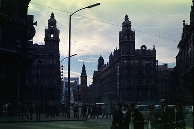 A bustling city street scene from the 1970s. Grand, ornate architecture dominates the view, likely a central European city. P...
