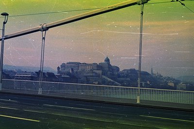 A vintage color photograph shows Budapest, Hungary, with Buda Castle prominently featured atop a hill, framed by a bridge’s s...