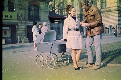 A stylish couple stands near an antique pram on a city street. The woman wears a belted coat dress; the man, a brown leather ...