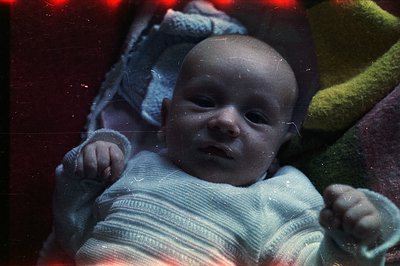 A baby, wrapped in a textured cream sweater, is centered in the frame. The image displays the characteristic color cast and v...