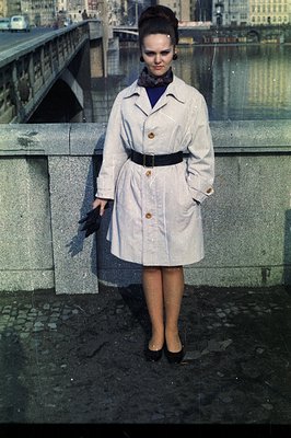 Young woman in a belted trench coat and dark dress stands on a stone bridge overlooking a river. Likely 1960s fashion, eviden...