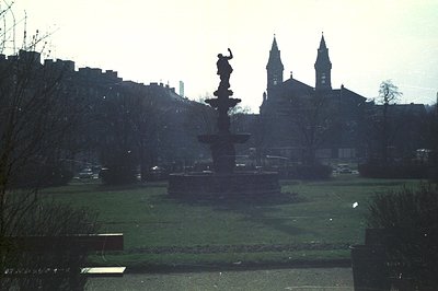A formal garden with a tiered fountain and statue dominates the view. Twin church spires rise in the background above a dense...