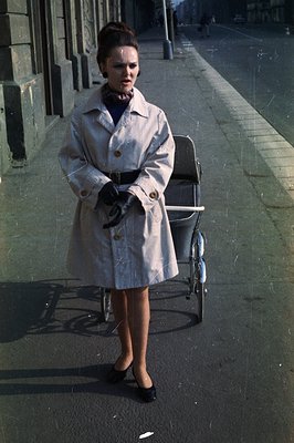 A stylish woman in a tan trench coat & dark dress stands next to a vintage stroller on a city street. Black gloves and a patt...