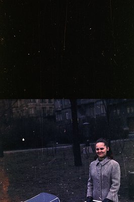 A young woman stands outdoors, posed near a park or garden. She wears a grey coat and gloves, her hair styled in a bob. Rain ...