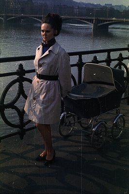A young woman in a belted trench coat and dark turtleneck stands beside a vintage pram overlooking a river and bridge. Likely...