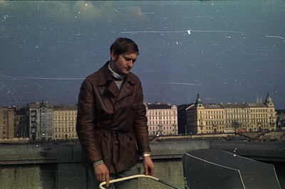 A man in a brown leather trench coat and turtleneck stands on a rooftop, overlooking a city skyline. Likely 1970s, based on s...