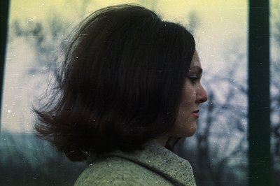 A young woman in profile, framed by a window with snowy tree branches visible beyond. She wears a light-colored knit sweater ...