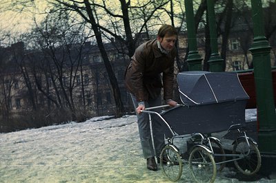 A man in a corduroy jacket pushes a vintage pram across a snowy park. Architectural backdrop suggests a European city, possib...